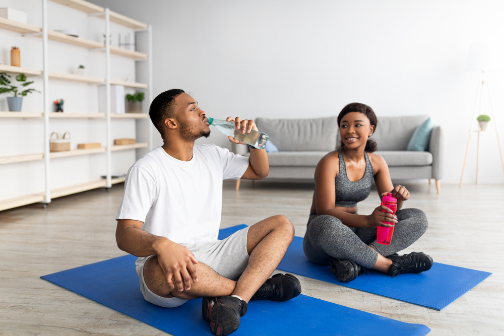 Athletic black couple in sportswear sitting on yoga mats, drinking water from bottles after domestic training, indoors